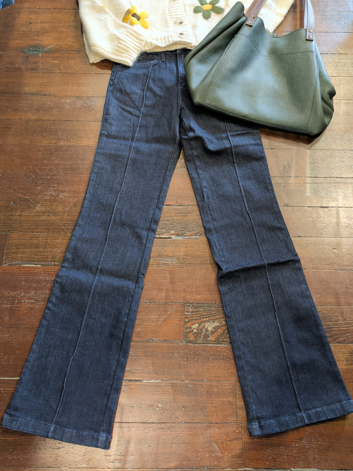 Wrangler Bespoke Camryn pintuck bootcut jeans laid flat with a cream floral cardigan and green tote.