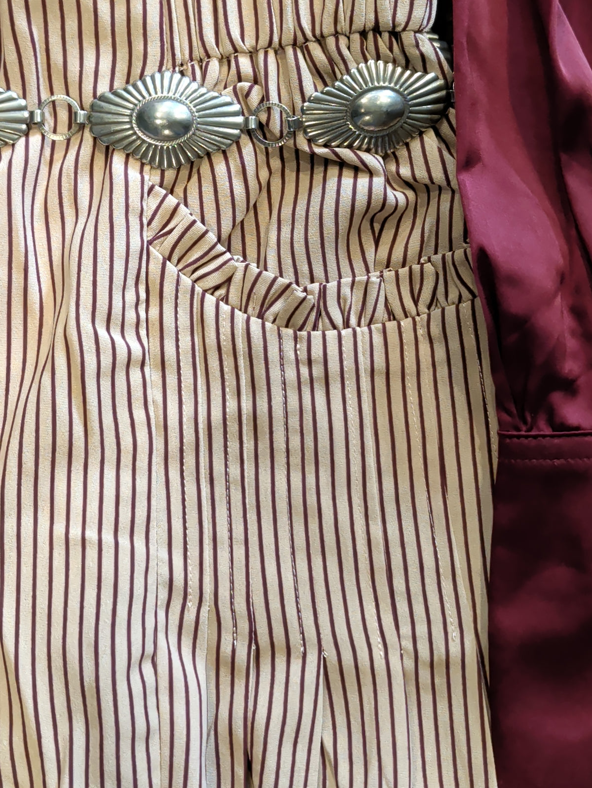 Close-up of tan palazzo pants showing burgundy pinstripes and a ruffle-trimmed patch pocket, styled with a silver-tone concho belt.
