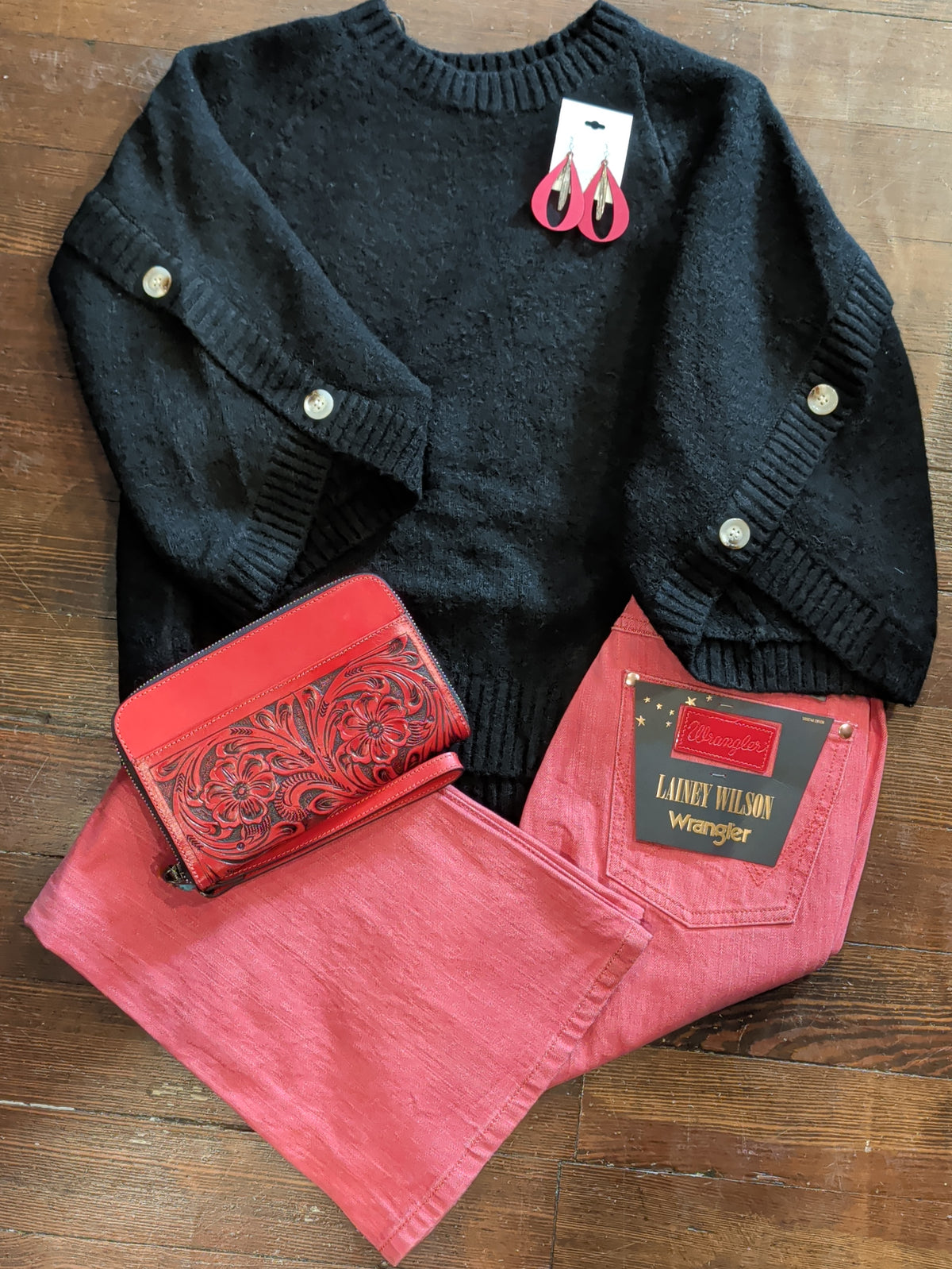 Black button-sleeve knit pullover styled flat with red Wrangler jeans, red teardrop earrings, and a red floral-tooled wallet.