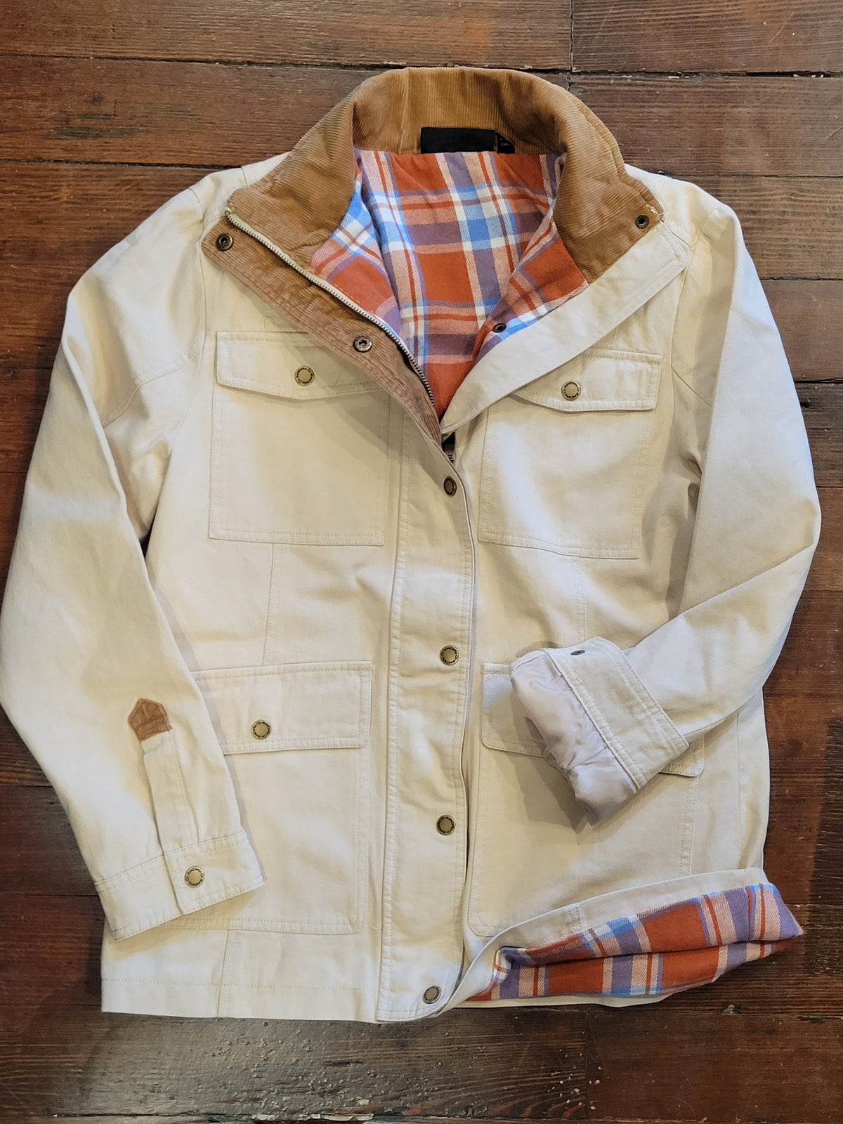 A cream-colored canvas Barn jacket/Coat with a brown corduroy collar, laid flat on a rustic wooden background. The jacket features a warm orange, blue, and white plaid lining visible inside the collar and cuffs.